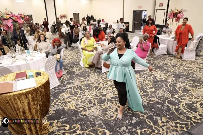 A woman in a mint dress sings into a microphone as she walks through a ballroom of women