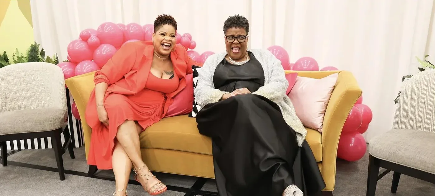 Two women laugh together on a gold sofa surrounded by pink balloons on stage